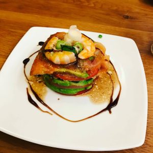 Auf einem viereckigen Teller angerichtet ein Turm aus verschiedenen runden Gemüsescheiben, darauf Garnelen. Kunstvoll dekoriert mit Lauch und Balsamico.