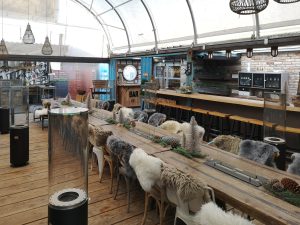 Gastraum mit langer Tafel, rustikale Holzmöbel, auf den Stühlen liegen Felle. Wintergartenbereich mit Heizvorrichtungen und Bar.
