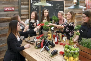 Sieben Frauen mit Kochschürzen trinken Sekt und unterhalten sich. Auf dem Tisch in Ihrer Mitte sind Auberginen, Paprika, Spirituosen, eine Kamera, Limetten und Küchenutensilien, wie ein Cocktailshaker und ein Kochmesser.