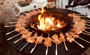 Rund um ein offenes Feuer sind marinierte Fleischspieße angerichtet